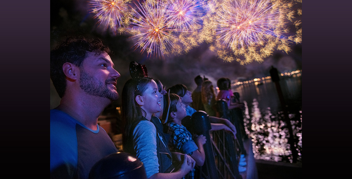 Guests stand against the railing at the World Showcase Lagoon at EPCOT. They stare out over the water with excited looks and smiles as they watch the show. Fireworks light up the sky above them.