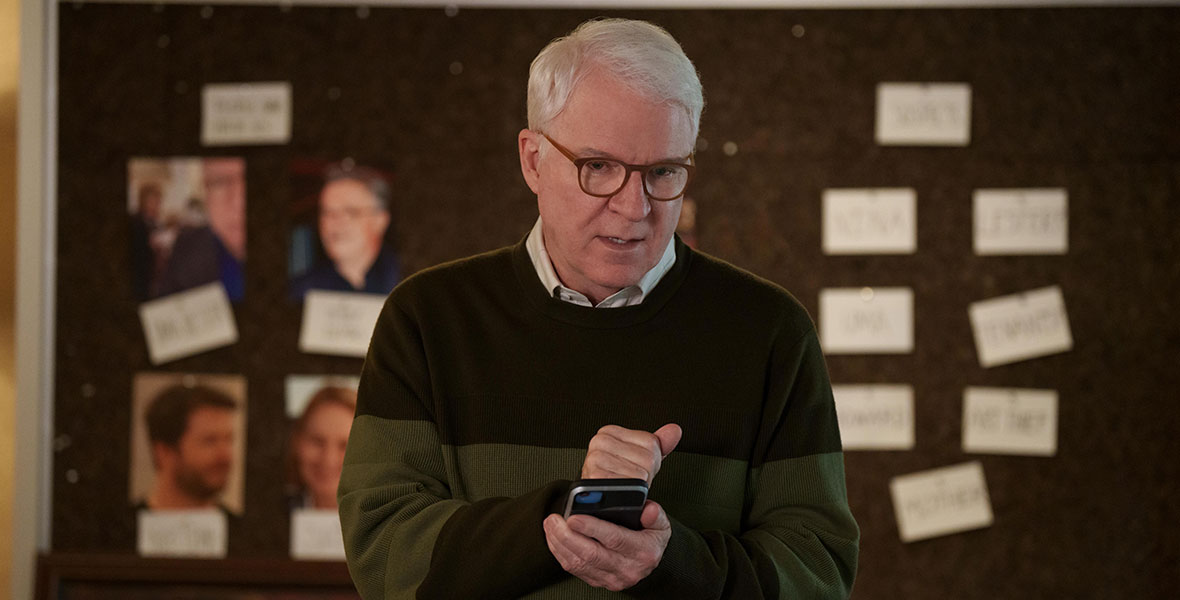 Disney Legend Steve Martin wears brown rimmed eyeglasses, a green and brown striped sweater, and a white dress shirt. He is holding a cell phone in his hands. Behind him is a corkboard with photos of potential murder suspects and other notes.
