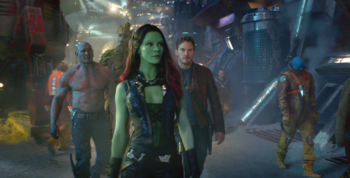 In a scene from Marvel Studios’ Guardians of the Galaxy, actors Zoe Saldaña as Gamora, Chris Pratt as Peter Quill, and Dave Bautista as Drax walk through a futuristic town. Saldaña has long black hair with a red ombre and green skin and wears a black, sleeveless top and black pants. Pratt wears a maroon leather jacket, black pants, and a gray T-shirt. Bautista is shirtless, exposing his gray skin and red markings, and wears black pants. Behind the group walks Groot, a large, tree-like humanoid.