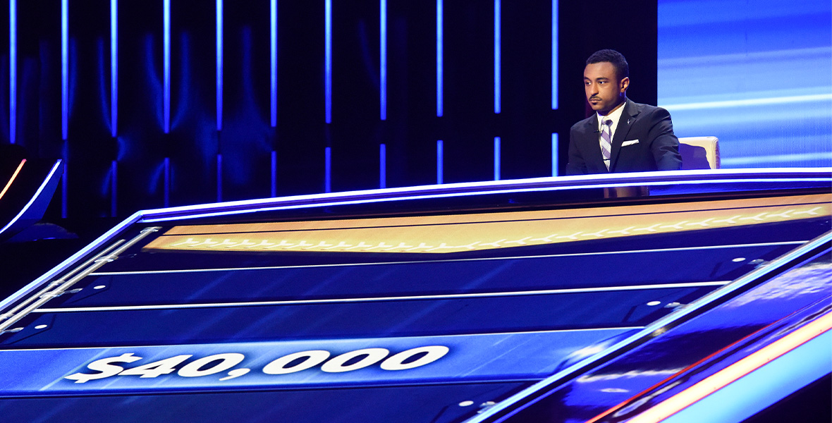 In a scene from ABC competition series The Chase, Brandon Blackwell sits above a large digital screen that reads “$40,000.” He wears a black blazer; a white button-down shirt; and a purple and silver-striped tie.
