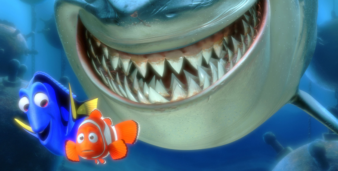 In a scene from Pixar’s Finding Nemo, a large great white shark smiles and looks at a Royal Blue Tang fish and Clownfish swimming next to it.