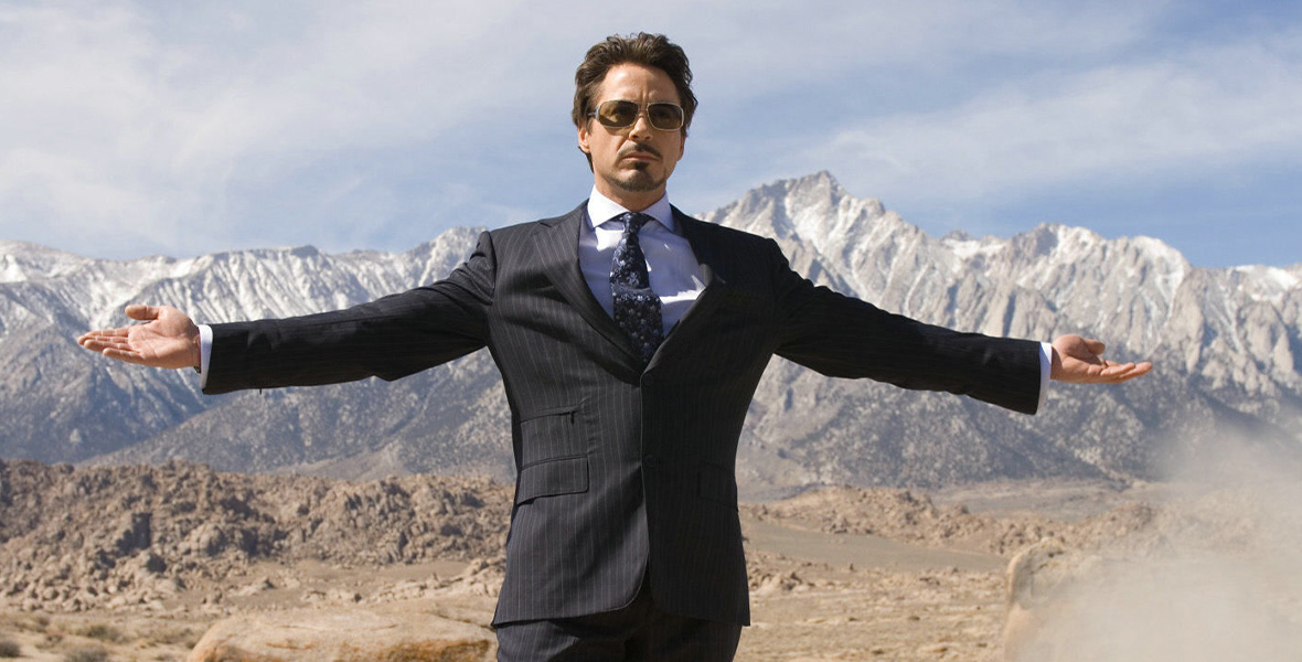 In a scene from Marvel Studios’ Iron Man, actor and Disney Legend Robert Downey Jr. portrays Tony Stark/Iron Man and wears a black suit with white pinstripes, a white button-down shirt, a floral print tie, and dark-lensed sunglasses. He extends both arms out as he stands in a barren desert.
