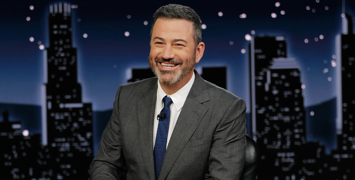 Host and comedian Jimmy Kimmel sits behind a wooden desk during an episode of Jimmy Kimmel Live!. He wears a gray suit, a white button-down shirt, and a navy tie. On the desk is a placard that reads “Jimmy Kimmel.”