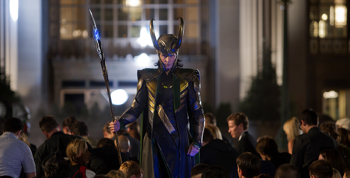 In a scene from Marvel’s The Avengers, actor Tom Hiddleston portrays Loki and wears a helmet with two horns, a long black and green leather coat, and black pants. He holds a staff in his right hand as he stands above a crowd of people.