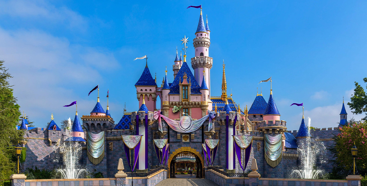 A rendering of Disneyland’s Sleeping Beauty Castle decorated for the Disney100 celebration. There are iridescent banners hanging from each turret and a large banner over the gateway reading “Disney 100”. Two fountains flank the drawbridge and the banners are being held up by Flora, Fauna, and Merrywether from Sleeping Beauty.