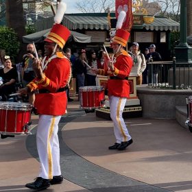 Four drummers dressed as toy soldiers are performing with Yamaha drums at Disney California Adventure Park at Disneyland Resort for the holidays in 2022. Their outfits are red with gold buttons and white pants with yellow stripes; their hats are red with yellow stripes. A crowd of onlookers surround them.