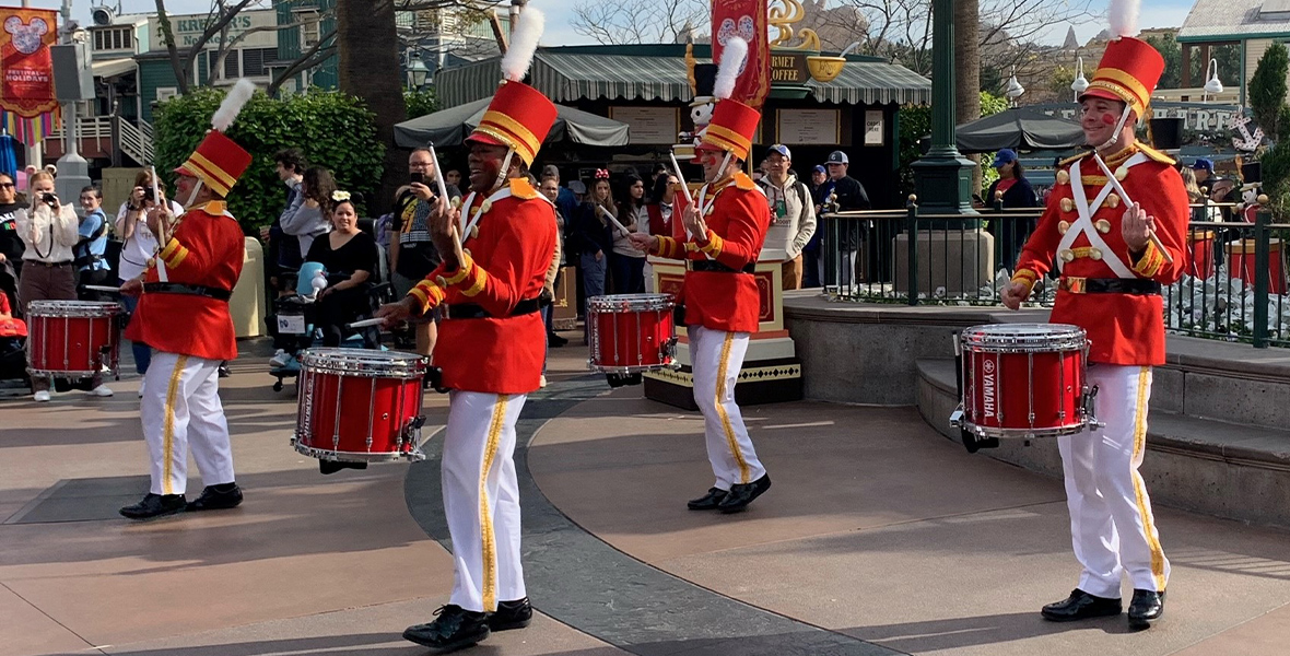 Four drummers dressed as toy soldiers are performing with Yamaha drums at Disney California Adventure Park at Disneyland Resort for the holidays in 2022. Their outfits are red with gold buttons and white pants with yellow stripes; their hats are red with yellow stripes. A crowd of onlookers surround them.
