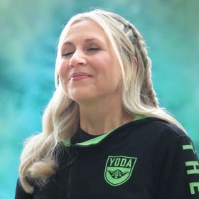 Ashley Eckstein faces towards the left with her eyes closed and a serene expression on her face. She stands against a lush green background, and we can see her from the chest up. She is wearing her blonde hair in a half-up braided style and has on a black, long sleeve top with a bright green Yoda emblem over her heart. There are additional bright green details around the neck and on the sleeves of the shirt.