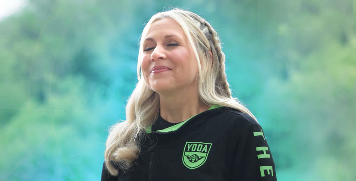 Ashley Eckstein faces towards the left with her eyes closed and a serene expression on her face. She stands against a lush green background, and we can see her from the chest up. She is wearing her blonde hair in a half-up braided style and has on a black, long sleeve top with a bright green Yoda emblem over her heart. There are additional bright green details around the neck and on the sleeves of the shirt.