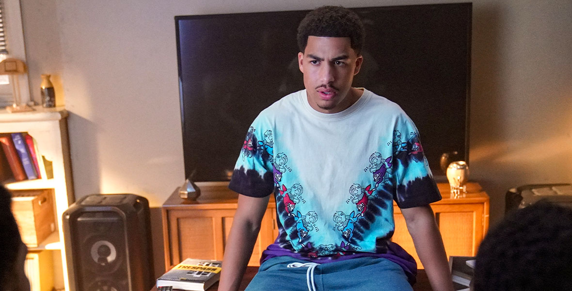 In a scene from Freeform series grown-ish, actor Marcus Scribner portrays Junior. Sitting on a wooden coffee table, he wears a colorful tie-dye shirt and blue sweatpants.