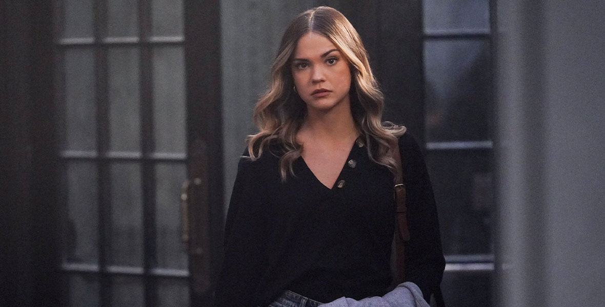 In a scene from Freeform series Good Trouble, actor Maia Mitchell portrays Callie Adams-Foster. She holds a gray coat over her arm, and wears a black, long-sleeved T-shirt and blue denim pants.