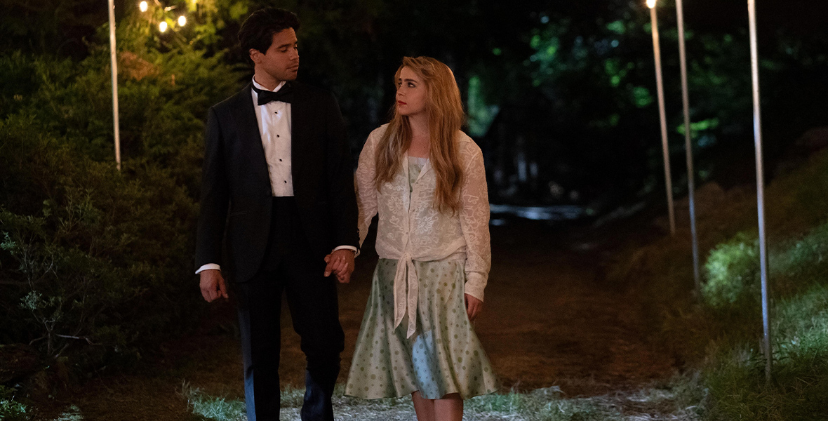 In a scene from Up Here, actors Carlos Valdes as Miguel and Mae Whitman as Lindsay walk hand in hand down a path surrounded by evergreen trees.