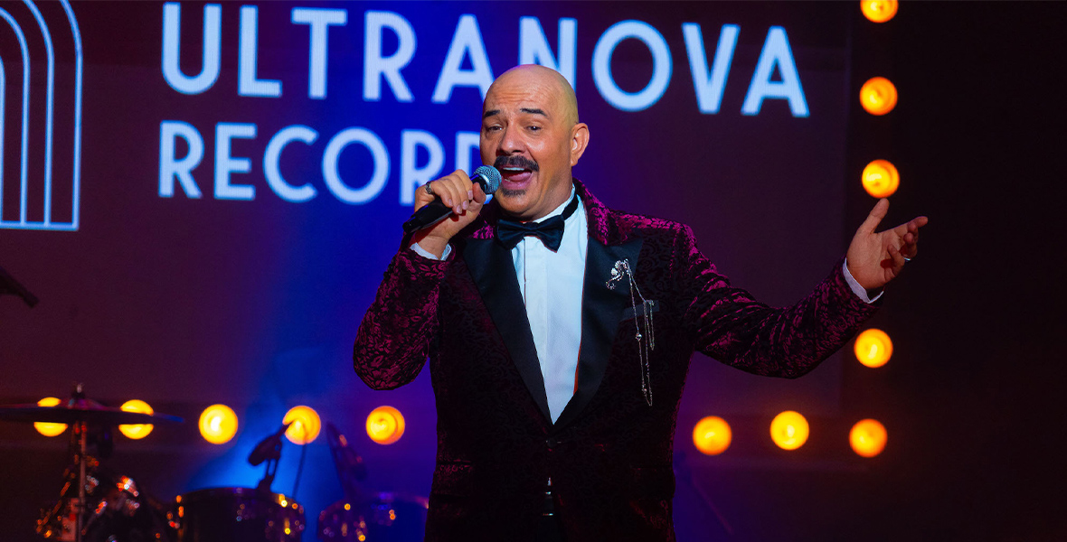 Principal Eduardo Kramer, played by Julián Arango, in The Low Tone Club, wears a purple and black suit, a white shirt, a silver brooch, and a black bowtie. He is bald and has a goatee. He stands centerstage and has a microphone in his right hand.