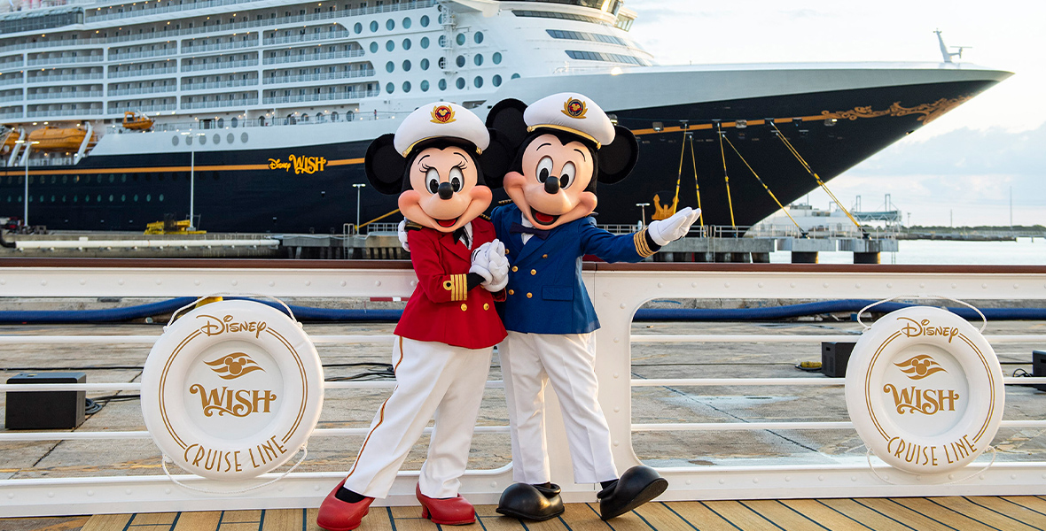 Captain Minnie, who wears a red jacket, a white hat, and white pants with red and gold stripes down the side, poses next to Captain Mickie, who wears a blue jacket, a white hat, and white pants. They stand on a dock in front of the Disney Wish, a massive cruise ship that showcases hundreds of staterooms, a smoke stack, and more.