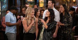 In a scene from Hulu Original series How I Met Your Father, actors Hilary Duff as Sophie and Francia Raisa as Valentina smile and point at each other in a crowded bar. Behind them are actors Tom Ainsley as Charlie and Tien Tran as Ellen, who both smile and look to their right. Duff wears a black dress under a sleeveless, cropped daisy top. Raisa wears a green, tie-dye mini dress and a black, leather blazer.