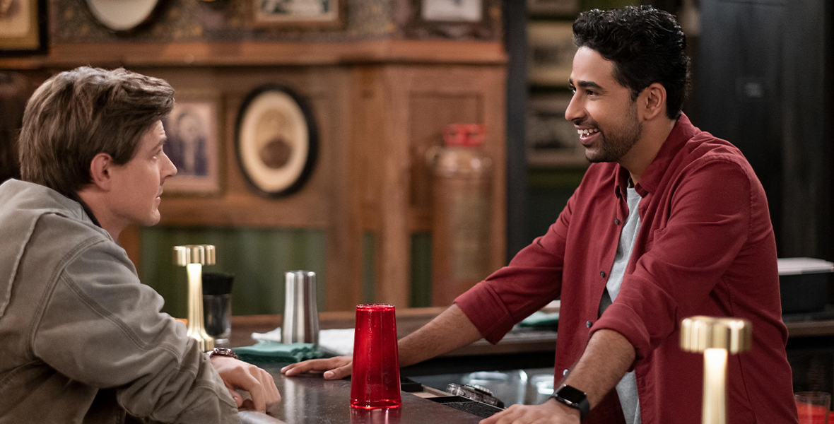 In a scene from Hulu Original series How I Met Your Father, actors Christopher Lowell as Jesse and Suraj Sharma as Sid talk to each other with a wooden bar top in between them. Lowell wears a tan jacket. Sharma wears a red button-down shirt and a gray T-shirt. A red plastic cup is upside down on the table.