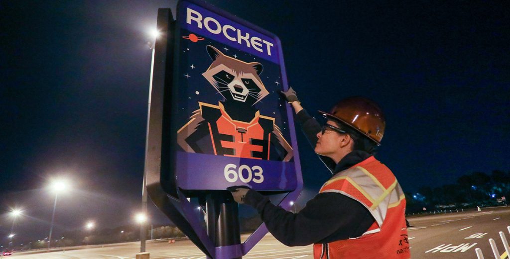 A cast member hangs a sign displaying the character Rocket from Guardians of the Galaxy in the parking lot at EPCOT.