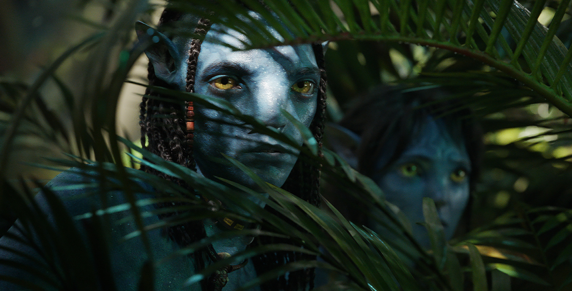 Two blue alien creatures, called Na’vi, peer through green leaves.