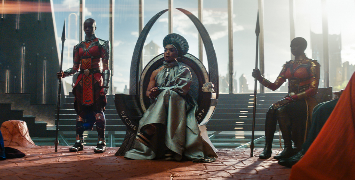 In a scene from Black Panther: Wakanda Forever, Queen Ramonda sits on a throne. She wears a silk one-sleeved dress and an ornate headpiece, and her hands are clasped in her lap. To her right is Ayo and to her left is Okoye, members of the Dora Milaje; they are wearing red, orange, yellow, and brown beaded uniforms with metal armor and holding spears in their right hands. The women are backlit by the bright sun.