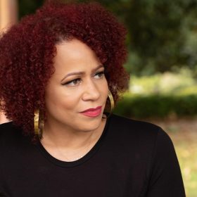 In a scene from The 1619 Project, Nikole Hannah-Jones sits outside; behind her are trees and bushes that are out of focus. Hannah-Jones has red, curly hair. She is wearing a simple black shirt, large gold hoop earrings, and a delicate gold necklace.