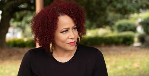 In a scene from The 1619 Project, Nikole Hannah-Jones sits outside; behind her are trees and bushes that are out of focus. Hannah-Jones has red, curly hair. She is wearing a simple black shirt, large gold hoop earrings, and a delicate gold necklace.
