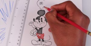 A hand is drawing Mickey Mouse on a piece of paper with a red pencil. The sketch of Mickey is colored in with watercolor paint. Mickey is wearing a thoughtful expression with an in-process lightbulb being drawn over his head. To his left is a white ruler with blue emphasis marks drawn next to it.