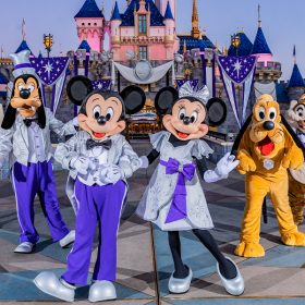 Daisy Duck, Donald Duck, Goofy, Mickey Mouse, Minnie Mouse, Pluto, Chip and Dale pose in front of Sleeping Beauty Castle at Disneyland park. All of the characters are dressed in platinum and purple outfits themed to Disney’s 100th anniversary.