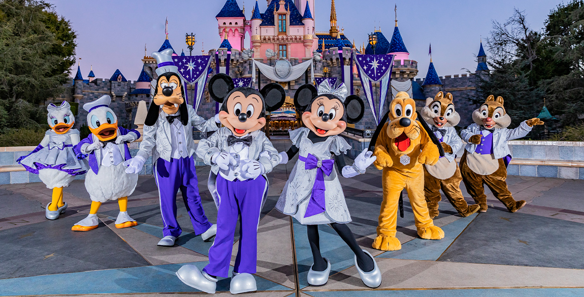 Daisy Duck, Donald Duck, Goofy, Mickey Mouse, Minnie Mouse, Pluto, Chip and Dale pose in front of Sleeping Beauty Castle at Disneyland park. All of the characters are dressed in platinum and purple outfits themed to Disney’s 100th anniversary.