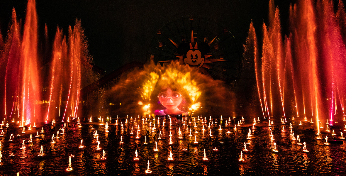 A body of water with fountains and water screens colored by bright red and orange lights. On the water screen is an image of Mirabel Madrigal from the film Encanto. Behind the screen, the Mickey Mouse face on the Pixar Pal-Around can be seen.