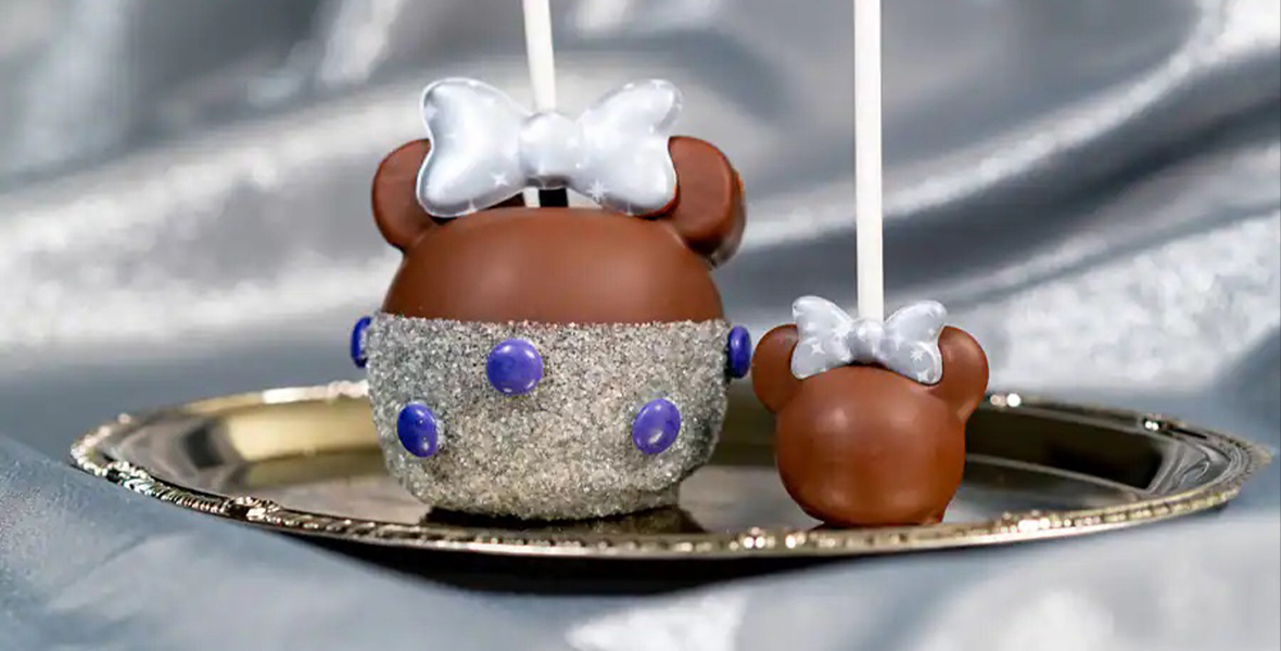 A chocolate-covered candy apple designed to look like Minnie Mouse wearing a sparkling dress. A smaller, all-chocolate candy apple Minnie sits next to it. Both are on a platinum platter set against a silver cloth backdrop.