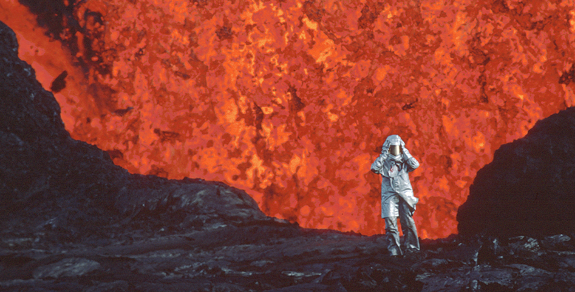 Maurice Krafft wears a full-bodied silver suit and puts his hands on his head. He is walking on hardened black volcanic rock as fiery orange lava erupts behind him.