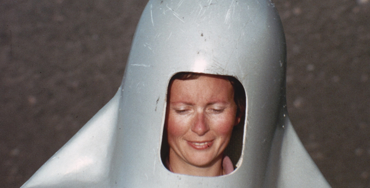 Katia Krafft wears a metal helmet-like contraption. The metal has scratch marks and scuff marks all over. Krafft’s face is visible through a rectangular cut-out.