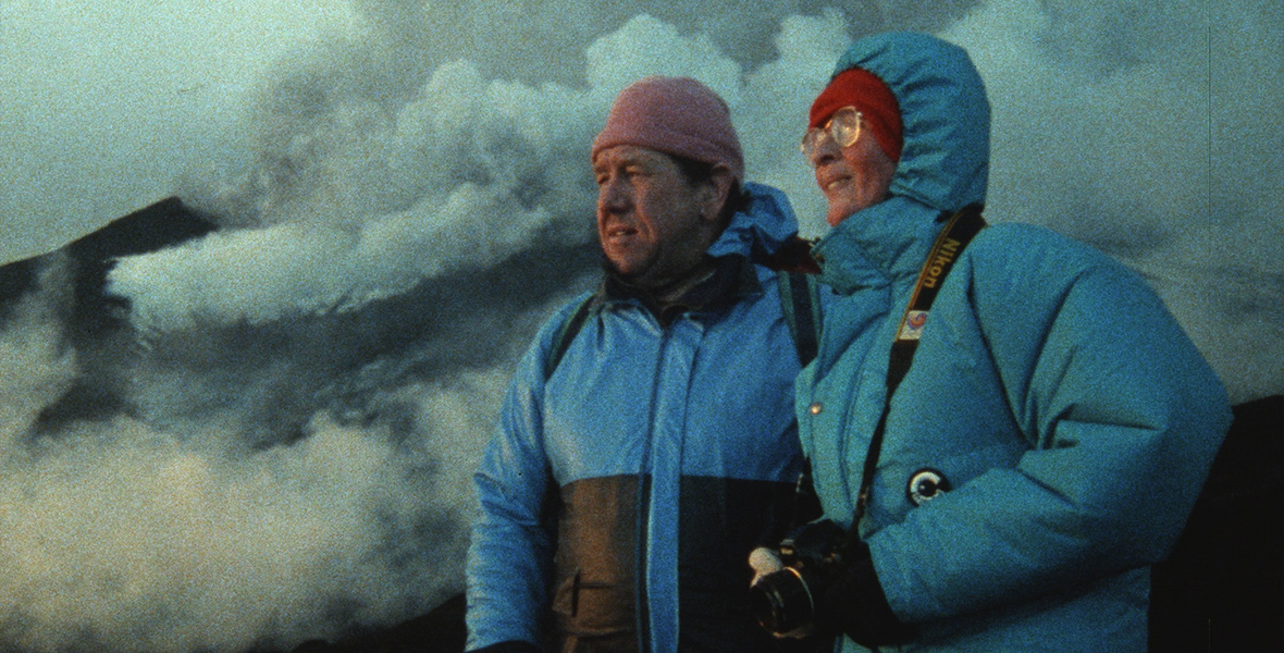 Maurice Krafft (left) and Katia Krafft (right) wear blue coats; Maurice’s coat has black color blocking from the ribs down. Maurice is also wearing a pink hat and carrying a green and black backpack. Katie is wearing a red hat and large glasses and is carrying a Nikon camera around her neck. Behind them are giant plumes of smoke.