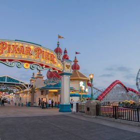 A view of Pixar Pier at dusk. The Pixar Pier marquee is lit up on the left side, paving the way to a lit archway that is boxed in by Knick’s Knacks and Lamplight Lounge. On the right is the water with the red tunnels of Incredicoaster behind it. Pixar Pal-A-Round is right next door, an enormous Ferris wheel with Mickey Mouse’s face at the center.