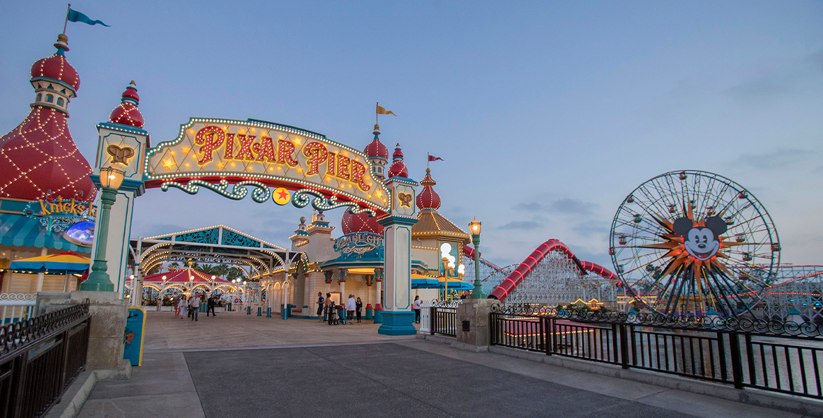 A view of Pixar Pier at dusk. The Pixar Pier marquee is lit up on the left side, paving the way to a lit archway that is boxed in by Knick’s Knacks and Lamplight Lounge. On the right is the water with the red tunnels of Incredicoaster behind it. Pixar Pal-A-Round is right next door, an enormous Ferris wheel with Mickey Mouse’s face at the center.