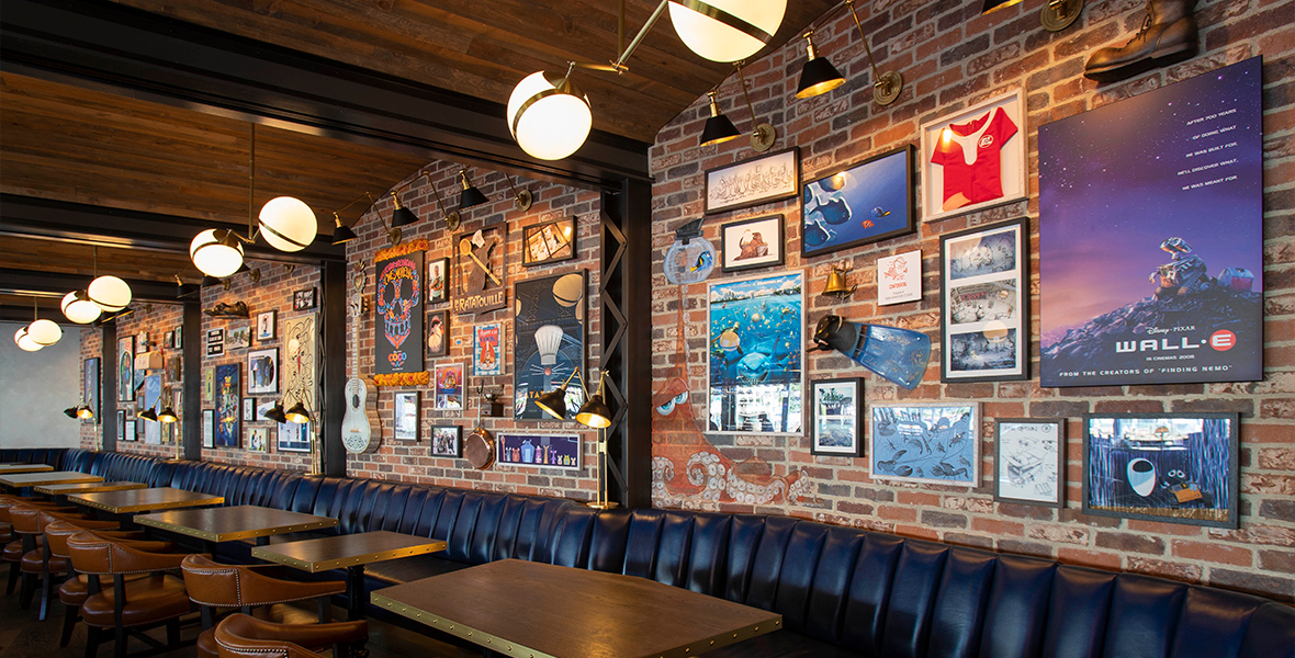 A view of Lamplight Lounge’s interior. A row of tables is pressed between a black banquette and a row of leather chairs. Pixar artwork and memorabilia, including the Finding Nemo film poster and the guitar from Coco, hang on the brick wall that the banquette leans against. Several desk-style lamps hang at the top.