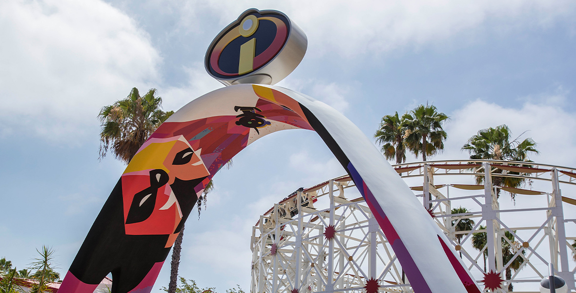 A view of the Incredibles-themed entrance just before Incredicoaster. The archway is Elastigirl stretching from one side to the other, with the rest of the Parr family painted on one side. The classic letter “I” logo sits atop the arch. In the background is the white rails of one of the coaster’s turns and the start of the queue.