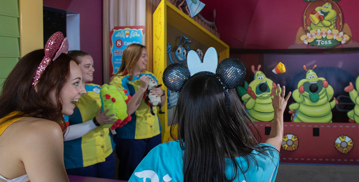 Two girls wearing mouse ears play Heimlich’s Candy Corn Toss. One, wearing a blue shirt, tosses candy corn toward one of three Heimlichs, all of whom have mouths open. On the side, two smiling cast members each hold a stuffed prize—one ladybug and one caterpillar—as they watch the candy corn midair.