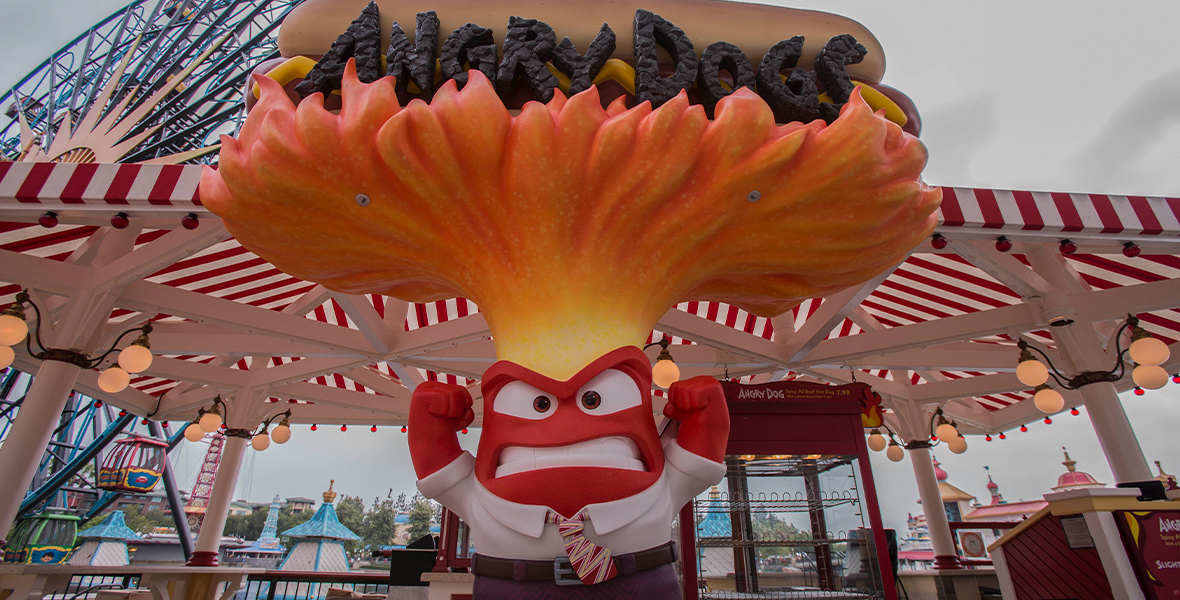A photo of the front of the Angry Dogs snack booth. A large figure of Anger, a bright-red emotion from Inside Out, wears a white collared shirt, red tie, belt, and dark trousers. His fists are thrown in the air, face enraged, as flames fly from the top of his head. Atop the horizontal flames is a hot dog, with “Angry Dogs” in black lettering across it.
