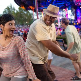 In the audience of an outdoor concert, a woman and man dance side-by-side as they look at each other, smiling. Two other couples dance in the aisle near them while a crowd sits on benches leading up to the main stage. In the background, the blurry stage is lit up in blues and pinks.