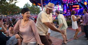 In the audience of an outdoor concert, a woman and man dance side-by-side as they look at each other, smiling. Two other couples dance in the aisle near them while a crowd sits on benches leading up to the main stage. In the background, the blurry stage is lit up in blues and pinks.