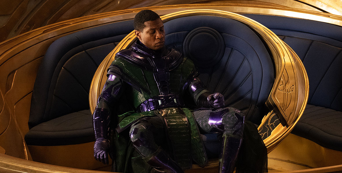 Kang the Conqueror, played by Jonathan Majors, wears a green and purple metallic comic book costume. He sits in a gold, circular chair in a gold room.