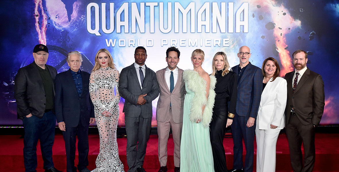 The cast and crew of Ant-Man and The Wasp: Quantumania on the red carpet in front of a large photo backdrop featuring the purple and pink swirls of the Quantum Realm. From left to right are Kevin Feige, Michael Douglas, Kathryn Newton, Jonathan Majors, Paul Rudd, Evangeline Lilly, Michelle Pfeiffer, Peyton Reed, Victoria Alonso, and Stephen Broussard.