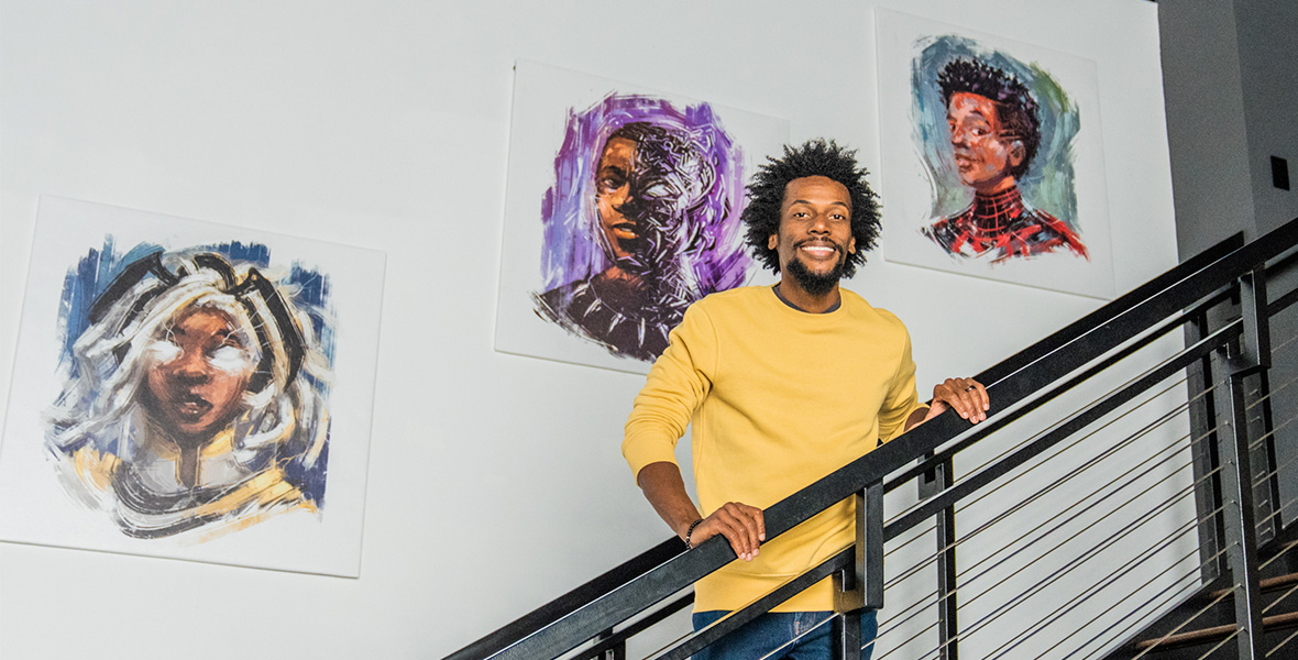 Artist Nikkolas Smith stands in the middle of a staircase, smiling at the camera. On the grey wall behind him are three Marvel Super Hero portraits that Smith created: Black Panther, Storm, and Miles Morales—all depicted in a colorful and sketchy style.