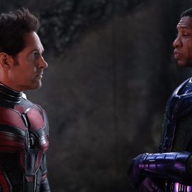 In a scene from Ant-Man and The Wasp: Quantumania, Scott Lang/Ant-Man (left) stands opposite Kang the Conqueror (right). Ant-Man is wearing a black and red suit; his helmet is off. Kang is wearing a purple suit with a green cape; his helmet is off.