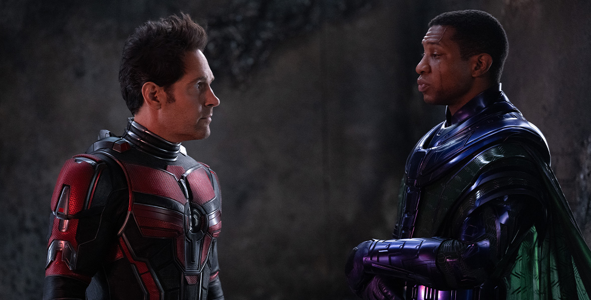 In a scene from Ant-Man and The Wasp: Quantumania, Scott Lang/Ant-Man (left) stands opposite Kang the Conqueror (right). Ant-Man is wearing a black and red suit; his helmet is off. Kang is wearing a purple suit with a green cape; his helmet is off.