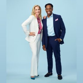Ali Wentworth wears a white suit, a pink top with a floral print, and blue pumps. She has her right arm on her hip and left arm on Dr. Adolph Brown’s right shoulder. Dr. Brown is wearing a navy suit, a blue shirt, a pink pocket square, and black dress shoes with gold buckles. They are smiling and posing against a blue backdrop.