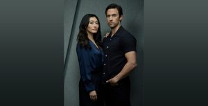 In a promotional photo shoot for The Company You Keep, Catherine Haena Kim stands to the right of Milo Ventimiglia. Kim is wearing a navy blue silk dress and rests her right hand on Ventimiglia’s right shoulder. Ventimiglia is wearing a black button-up shirt and black pants, and his left hand is in his pocket. Neither is smiling.