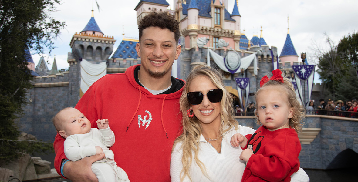 An image of MVP Patrick Mahomes and his family, standing in front of Sleeping Beauty Castle at Disneyland Park; he is holding his infant son Patrick “Bronze” Lavon Mahomes III; to the right of the elder Patrick is his wife Brittany, who is holding their toddler daughter Sterling Skye. The elder Patrick is wearing a red hoodie; Brittany is wearing a white long-sleeved shirt and sunglasses and has blond hair; Sterling Skye is wearing a red Minnie Mouse sweatshirt and has a red bow in her hair.