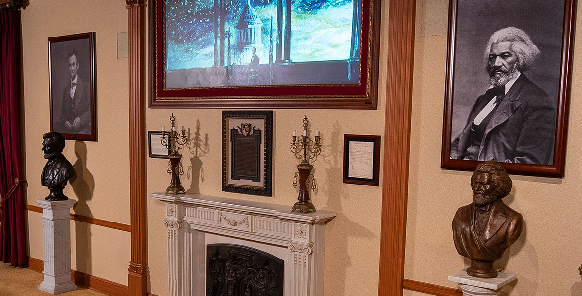 A photo of Main Street Opera House’s new exhibit, which takes up space on a cream-colored wall. Against the wall is a small, white fireplace mantle with two candelabras, one on each side. Above that is a certificate, and a large painting hangs above of a man sitting in front of a starry U.S. Capitol. To the mantle’s right is a bust of Abraham Lincoln and his portrait just above it. To the mantle’s left is a bust of Frederick Douglas and his portrait above it.